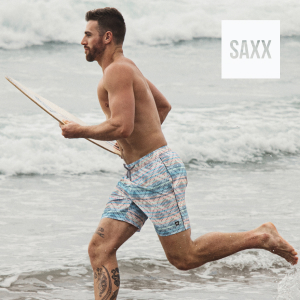 Man running on the beach with his surfboard, wearing geometric‑print swim shorts from SAXX.