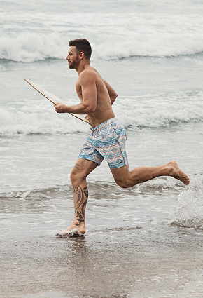 Man running on the beach in front of a surfboard and wearing geometric-pattern swim trunks from the brand Saxx. 